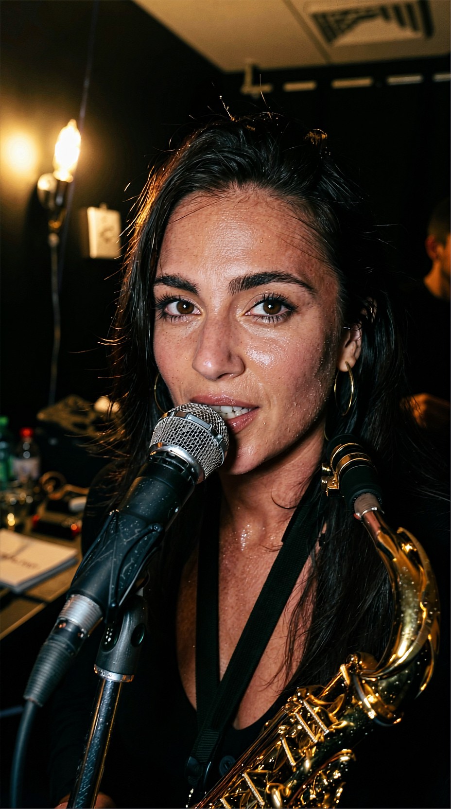 Close-up of a dark-haired woman with wet-looking skin and freckles singing into a microphone, a brass saxophone strap across her chest, warm bulb on the left wall, a man partly visible at the right edge